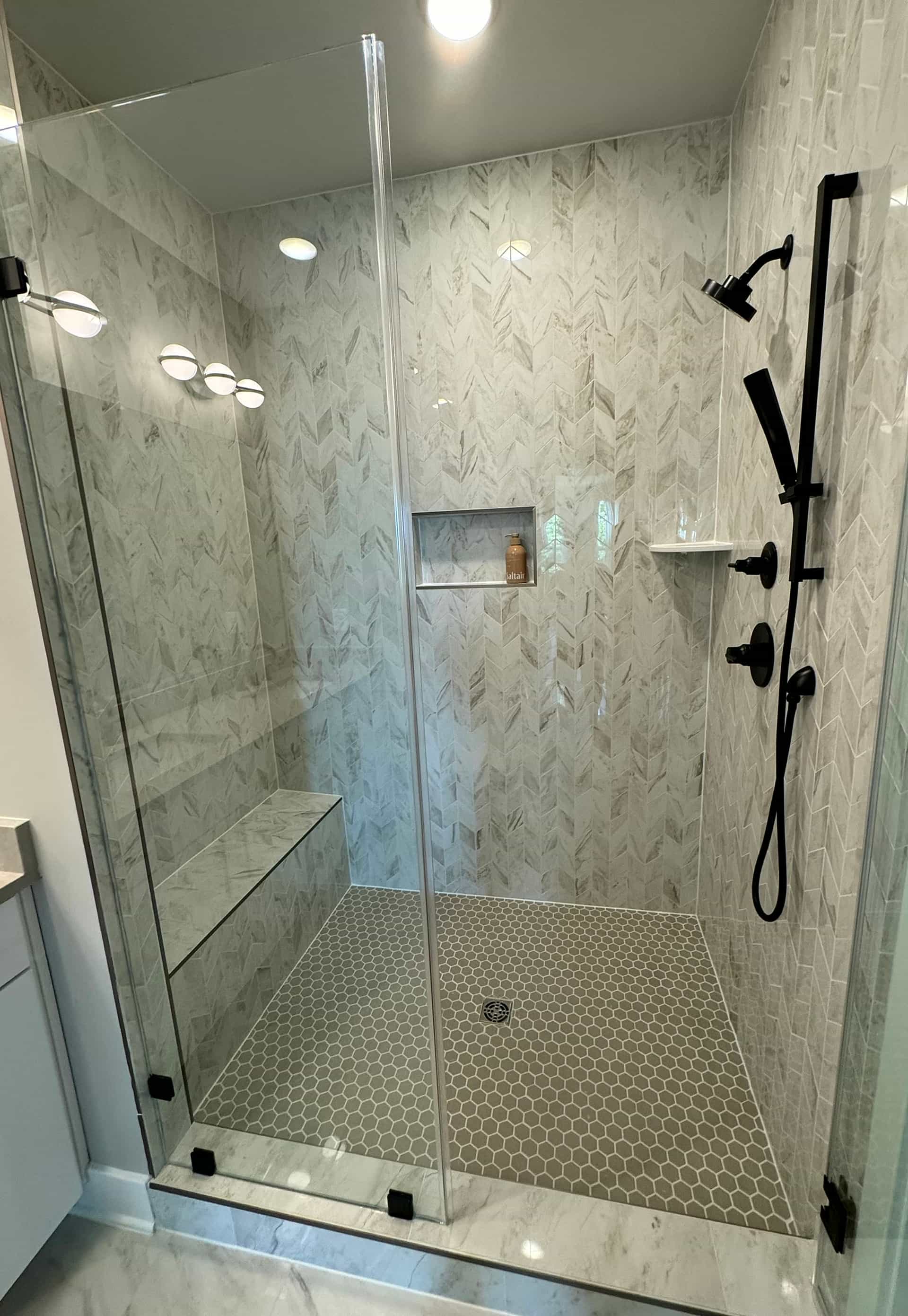 Floor-to-ceiling marble chevron shower with frameless glass, tiled bench, hex floor, and matte black fixtures