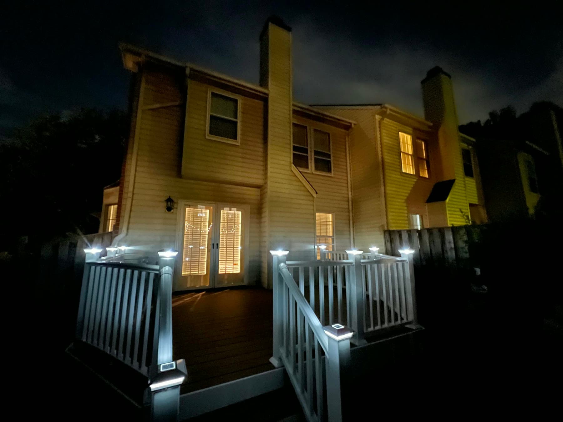 Composite deck at night with white vinyl railings and glowing post-cap lights below lit French doors